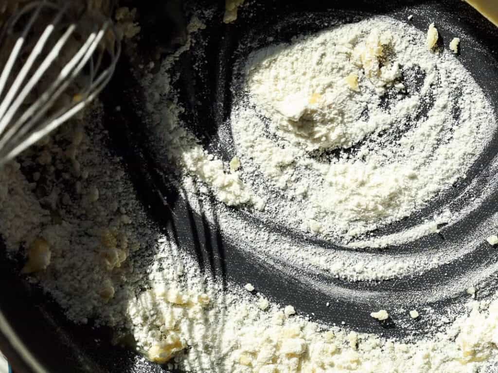 A close-up of a dusty black surface with flour and lumps scattered across it. A whisk sits partially submerged in the flour, with light creating shadows and contrasts in the flours texture.